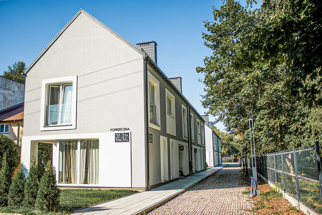 Nowoczesny szeregowiec na osiedlu Cichy Zakątek przy ulicy Poprzecznej, z jasną elewacją i dużymi oknami. Przed budynkiem rosną tuje, a obok znajduje się brukowana alejka prowadząca wzdłuż ogrodzenia z siatki. W tle widać kolejne segmenty domów w zabudowie szeregowej.